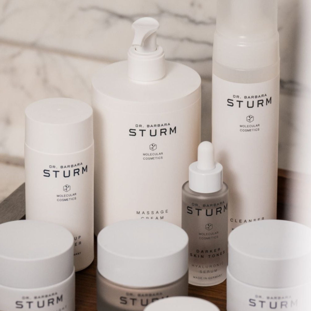 A collection of Dr. Barbara Sturm skincare products in white and frosted glass containers, arranged on a wooden tray against a marble background.
